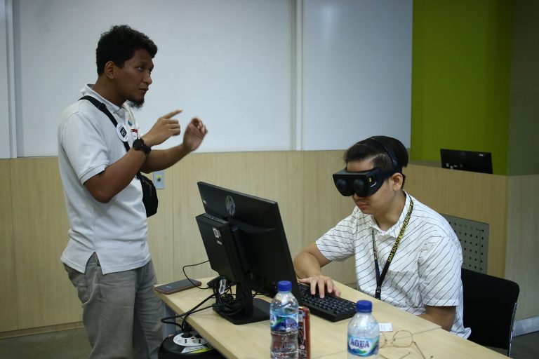 seorang pria teman netra sedang menjelaskan cara penggunaan pembaca layar atau screen reader kepada pria awas yang menggunakan kacamata simulasi tunanetra sambil mencoba pembaca layar di komputer