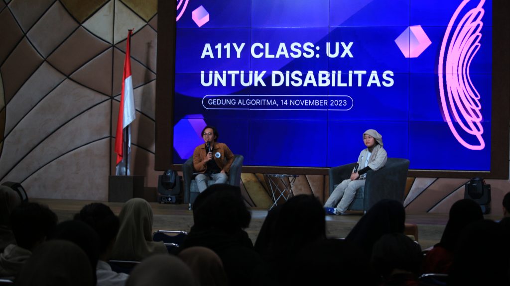 dua orang sedang duduk di hadapan para penonton. mc (kiri) memperkenalkan pembicara rahma. Di belakang mereka ada layar bertuliskan Accessibility atau alli class ux untuk disabilitas, egdung algoritma 14 November 2023