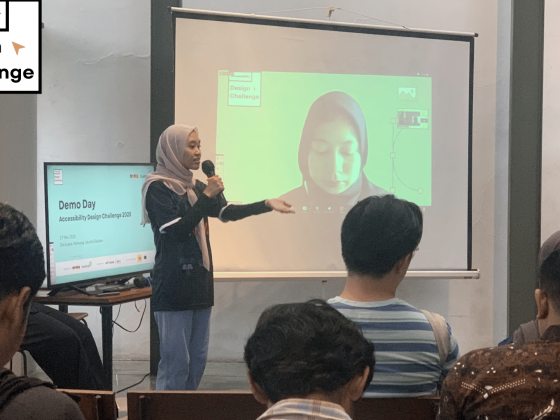 Foto suasana sebuah ruangan yang berisi sekumpulan orang duduk dan menghadiri event. Tampak seorang perempuan berhijab, mengenakan mic di depan audiens yang duduk, dengan membelakangi sebuah tv bertuliskan Demoday Accessibility design challenge, dan layar yang terdapat seorang perempuan bersiap untuk presentasi.